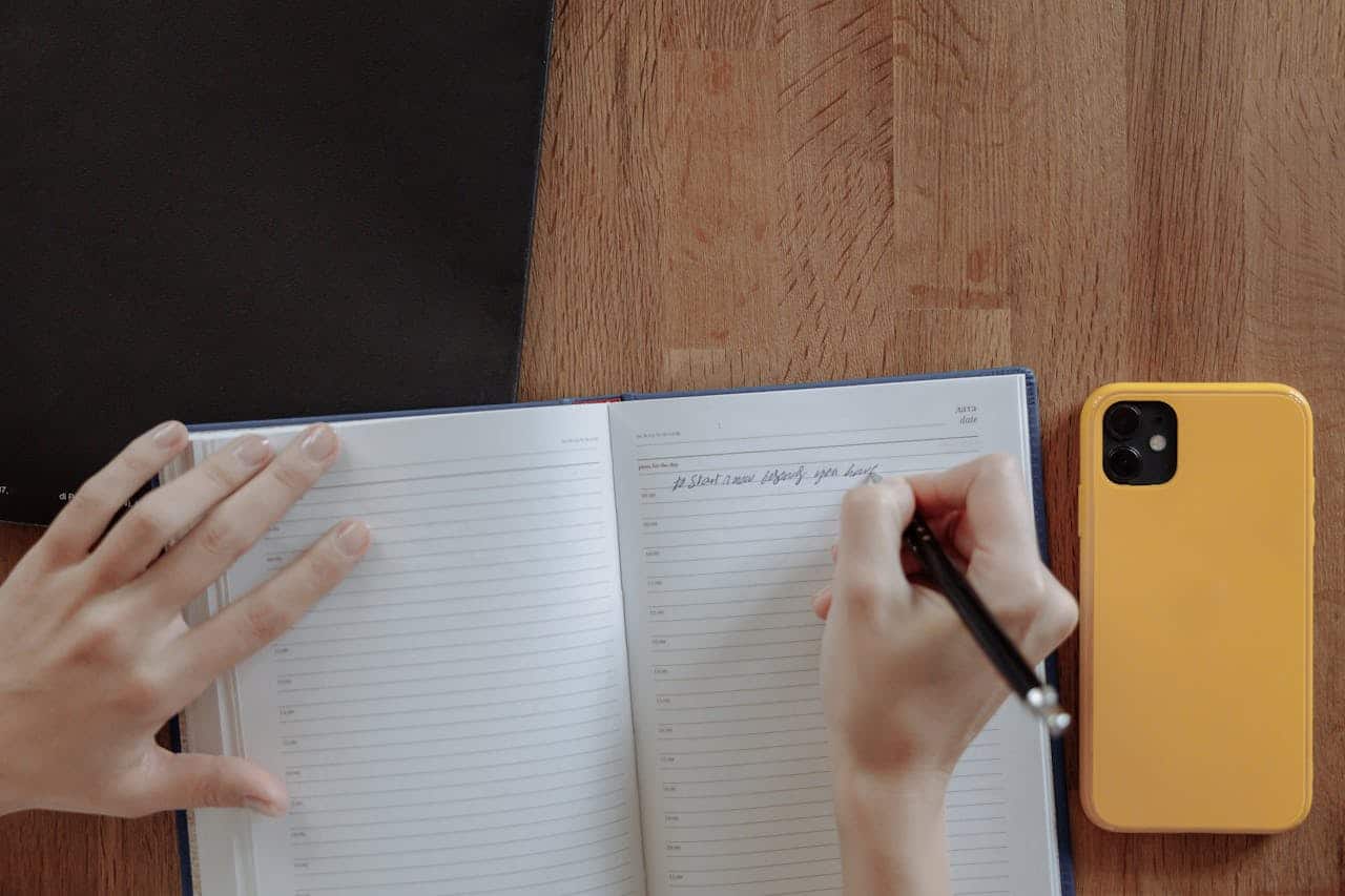 Hands writing in a notebook on a wooden table with a yellow smartphone nearby, promoting productivity.